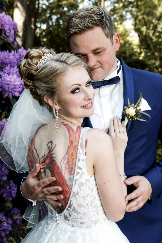 Hochzeitsfotograf Franz Fender: Die Braut trägt ein am Rücken tief ausgeschnittenes weißes Kleid. Das großflächige bunte Tattoo ist teils verdeckt von der Hand des Bräutigams, mit der er sie sanft an sich drückt. Sie blickt lächelnd über die Schulter in die Kamera. Sein Blick ruht auf ihr. Aufgenommen im Garten des Wilhelm-Busch-Museums, Hannover.